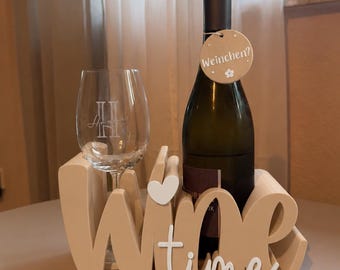 Könnte beinhalten: Ein beigefarbener Weinflaschenhalter in Form des Wortes Wine mit einem weißen Herzakzent. Ein klares Weinglas mit Monogramm steht im Halter neben einer dunklen Weinflasche. Ein rundes beiges Etikett mit dem Text Weinchen? hängt am Flaschenhals.