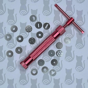 Könnte beinhalten: Ein rotes Metallwerkzeug mit einem Griff und einem T-förmigen Hebel. Das Werkzeug wird verwendet, um verschiedene Formen aus Ton oder Fondant zu erstellen. Es gibt 17 verschiedene Metallscheiben mit verschiedenen Formen und Mustern.