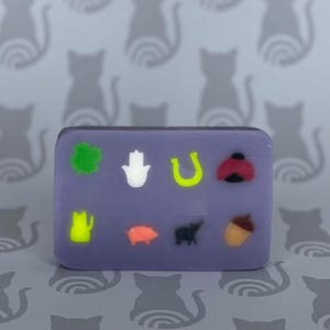 Könnte beinhalten: Eine violette Seifenstange mit verschiedenen geprägten Symbolen, darunter ein vierblättriges Kleeblatt, eine Hand, ein Hufeisen, ein Marienkäfer, ein Friedenszeichen, ein Schwein, ein Bär und eine Eichel.