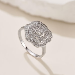 May include: A silver ring featuring a rose design. The rose is encrusted with small, clear gemstones, creating a sparkling effect. The ring has a delicate band and a detailed floral centerpiece, perfect for a special occasion.