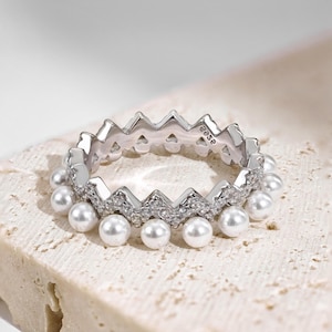 Può includere: Un anello d'argento con una fila di piccole perle bianche e un design a zig-zag con piccole pietre chiare. L'anello è contrassegnato con "925e".