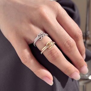 Op de afbeelding: Twee ringen, één zilverkleurig en één goudkleurig, elk met een knoopontwerp. De banden zijn bezet met kleine, heldere edelstenen. De ringen worden op een hand getoond, waarbij de sieraden worden uitgelicht.