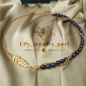 May include: Gold-toned necklace with dark blue pearls and a leaf design. The necklace is displayed on a satin fabric with a teal box in the background. The text "Lily_jewelry_perl" is visible.