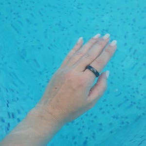 Könnte beinhalten: Ein schwarzer Ring an einer Hand in blauem Wasser. Der Ring ist ein einfaches Band. Das Wasser hat einen Welleneffekt, und die Hand befindet sich in der Mitte des Rahmens. Das Wasser scheint ein Schwimmbecken zu sein.