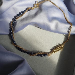 May include: A gold chain necklace with a gold leaf pendant and black pearls. The necklace is on a blue fabric background.