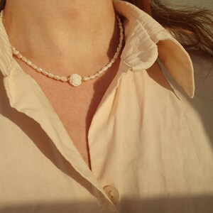 Puede incluir: Un delicado collar de perlas con un colgante central de rosa blanca. El collar está compuesto por pequeñas perlas irregulares y se lleva sobre una camisa de cuello claro.