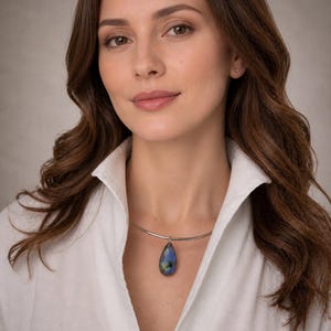 Op de afbeelding: Een vrouw draagt een wit overhemd en een zilveren ketting. De ketting heeft een druppelvormige hanger met een blauw-groene iriserende steen. De hanger contrasteert met het witte overhemd en de neutrale achtergrond.