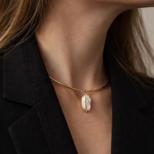 May include: A gold-toned necklace with a pendant featuring a large, irregular-shaped white pearl. The pearl has a textured surface and is suspended from a delicate gold chain. The necklace is worn against a black blazer.