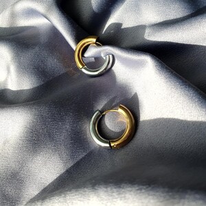 Puede incluir: Dos pendientes de aro bicolor, cada uno con un diseño dorado y plateado. Los pendientes se exhiben sobre una tela gris y sedosa. Diseño sencillo y moderno.