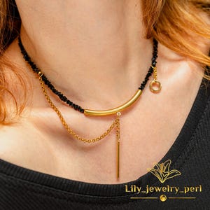 May include: A gold-toned necklace with a black beaded strand. The necklace features a curved bar pendant, a dangling chain, and a small circular charm. The necklace is worn on a person's neck. The text "Lily_jewelry_perl" is visible.