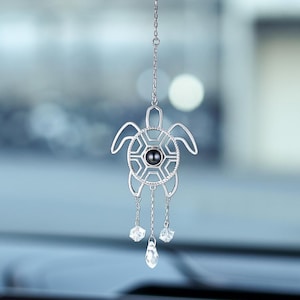 May include: A silver-coloured car charm shaped like a turtle, suspended from a chain. The turtle features a black centre and three clear crystal dangles. The charm hangs in front of a car's interior.