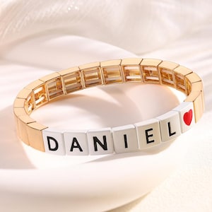 May include: Gold-toned bracelet with the name "DANIEL" spelled out in black letters on white blocks. A small red heart is at the end of the name. The bracelet is on a white surface.