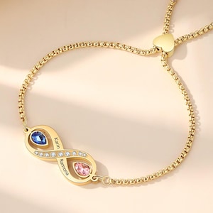 Puede incluir: Pulsera dorada con símbolo de infinito y cierre en forma de corazón. El símbolo del infinito presenta una piedra preciosa azul y rosa, con los nombres "Karin" y "Ramona" grabados. La pulsera está sobre una superficie beige claro.