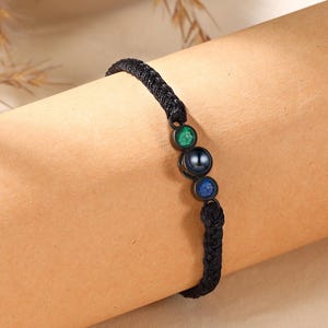 May include: Black braided bracelet with a series of colorful beads. The bracelet features a green, a dark gray, and a blue bead. The bracelet is worn on a person's arm.