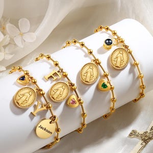 May include: Gold charm bracelets with religious medallions, initial letters, and a charm engraved with the name "Jackson." The bracelets feature small, round beads and are adorned with colourful gemstones. The bracelets are displayed on a white surface.