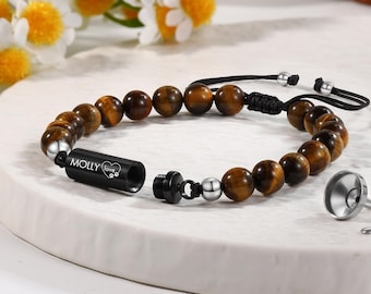 May include: A beaded bracelet with brown tiger eye stones and a black cylinder pendant engraved with "MOLLY love". Includes a small silver funnel and tool. The bracelet is on a white surface with flowers in the background.