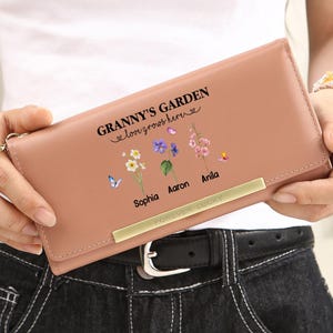 May include: A blush-colored wallet with the text "GRANNY'S GARDEN love grows here" and floral illustrations. The wallet has a gold-colored metal bar and the words "FOREVER LUCKY" are printed on the bottom. The wallet is held by a person.