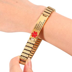 Może przedstawiać: Złota bransoletka medyczna z czerwonym symbolem medycznym i wygrawerowanym tekstem. Tekst brzmi "Sonja Miles, Lymphedema Alert, No BP/IV left arm, A Miles ICE 409 893 2426". Bransoletka jest pokazana na nadgarstku.