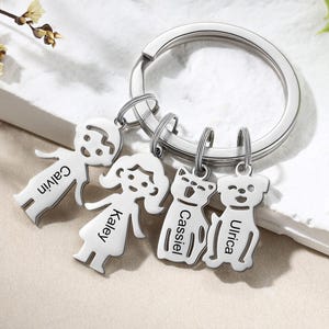 Può includere: Un portachiavi argentato con quattro ciondoli personalizzati. I ciondoli includono due figure umane e due figure di cane, ognuna incisa con un nome: Calvin, Kaley, Cassiel e Ulrica. Il portachiavi è su una superficie bianca.