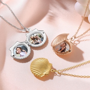 Puede incluir: Collares con medallones personalizados en tonos plata y oro. Los medallones están abiertos, mostrando fotografías. Uno tiene forma de concha. Los collares se exhiben sobre una superficie rosa suave.