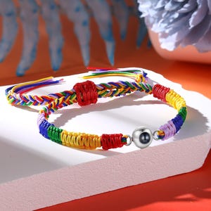 Puede incluir: Dos pulseras trenzadas coloridas. Una pulsera presenta un nudo cuadrado rojo y un diseño trenzado con hilos de colores del arcoíris. La otra pulsera tiene una cuenta plateada y está hecha de hilos de colores del arcoíris.