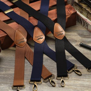 May include: A collection of suspenders in various colors: brown, navy blue, and black. Each suspender features a leather patch with the word "Spoon" and a mustache graphic. The suspenders have gold-colored metal clasps and adjusters.