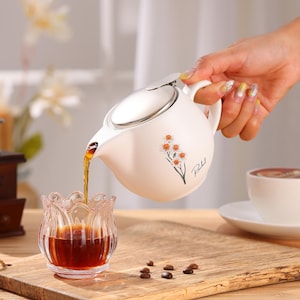 May include: A white ceramic teapot with a silver lid is pouring a dark liquid into a clear glass. The teapot has a floral design and a handle. A cup of coffee sits on a white saucer. The scene is set on a wooden surface.