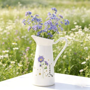 Puede incluir: Una jarra de cerámica blanca con asa, llena de flores azules. La jarra tiene un diseño floral y el nombre "Janice" en escritura cursiva. Puntos dorados acentúan el diseño floral. La jarra está sobre una superficie blanca con un fondo borroso de flores.