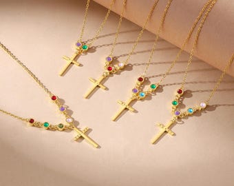 Puede incluir: Varios collares de cruz dorados con piedras preciosas de colores. Cada collar presenta una cadena delicada y un colgante de cruz. Las piedras preciosas son de varios colores, incluyendo rojo, verde, morado y amarillo. Los collares se exhiben sobre una superficie marrón claro.