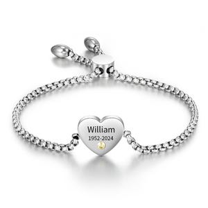 Op de afbeelding: Zilverkleurige armband met een hartvormige bedel gegraveerd met de naam "William" en de jaren "1952-2024". Een kleine, gele edelsteen is gezet aan de onderkant van het hart. De armband heeft een verstelbare schuifsluiting.