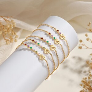 May include: A set of five gold-toned chain bracelets, each featuring a small heart-shaped charm and a series of multi-colored round gemstones. The bracelets are displayed on a white cylindrical stand, showcasing delicate links and vibrant stones.