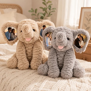 Peut inclure: Deux éléphants en peluche, l'un beige et l'autre gris, avec une fourrure douce et texturée, de grandes oreilles et une petite langue rose. Chaque éléphant présente une photographie circulaire imprimée à l'intérieur des oreilles, montrant un couple.