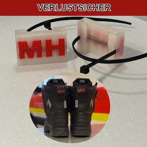 Könnte beinhalten: Zwei transparente Kunststoffanhänger mit den roten Buchstaben "MH", befestigt mit schwarzen Kabelbindern. Der Text "VERLUSTSICHER" steht oben. Darunter sind zwei schwarze Stiefel mit "MH" auf der Rückseite zu sehen.
