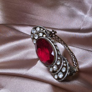 Könnte beinhalten: Ein verzierter Armreif mit einem großen, ovalen, rubinroten Edelstein als Mittelpunkt. Der Armreif weist aufwendige Details mit kleineren, klaren Edelsteinen und einer bronzefarbenen Metalleinfassung auf. Der Hintergrund ist ein Satin-Stoff.