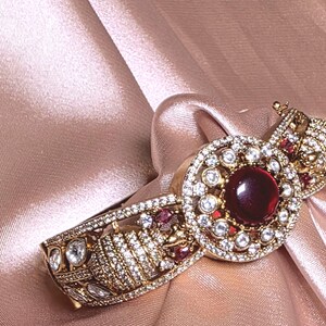 以下が含まれることがあります： クリアと赤の宝石をあしらった、精巧なディテールが施されたゴールドトーンのブレスレット。ブレスレットの中央には、小さなクリアストーンに囲まれた大きな赤い石を使った円形デザインがあります。背景は柔らかいピンク色の生地です。