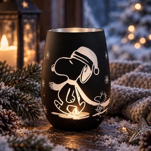 May include: A matte black cylindrical candle holder featuring a white silhouette graphic of Snoopy wearing a Santa hat and a scarf, accompanied by Woodstock. The design includes small snowflake accents and a cutout at the base for a tea light.