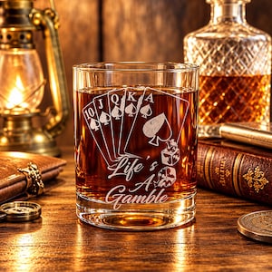 May include: A clear glass tumbler filled with amber liquid, featuring an etched design of a royal flush in spades, two dice, and the text Life's A Gamble. The glass has a thick base and straight sides, showcasing the detailed white engraving.