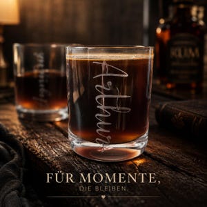 Könnte beinhalten: Zwei gravierte Whiskygläser gefüllt mit einer dunklen Flüssigkeit. Das vordere Glas ist mit dem Namen "Arthur" graviert. Die Gläser stehen auf einer Holzoberfläche. Das Bild enthält eine Rumflasche und den Text "FÜR MOMENTE, DIE BLEIBEN."