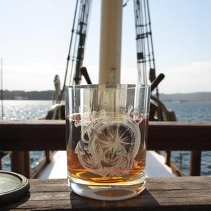 May include: A clear glass with an etched design of an anchor and compass on a wooden surface. The glass is filled with a dark amber liquid.
