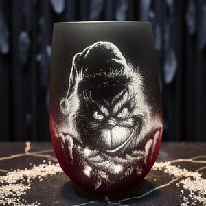 Könnte beinhalten: Ein stieloses Weinglas mit einer mattierten schwarzen Außenseite, die in eine tiefrote Basis übergeht. Das Glas zeigt eine detaillierte, weiß geätzte Illustration einer Figur mit Weihnachtsmütze und einem schelmischen Gesichtsausdruck.