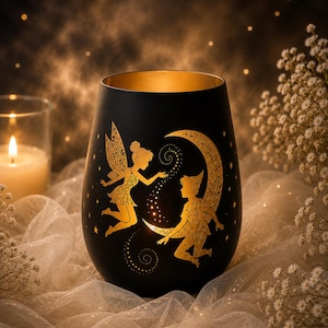 Könnte beinhalten: Ein mattschwarzer, zylindrischer Kerzenhalter mit einer goldfarbenen Silhouette einer Fee und eines Jungen, der auf einer Mondsichel sitzt. Das Design zeigt filigrane Muster auf den Figuren und dem Mond sowie ein warmes Gold-Finish im Inneren.