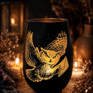 Könnte beinhalten: Ein mattschwarzes Glasgefäß mit einer goldfarbenen, geätzten Illustration einer Eule im Flug, die einen versiegelten Umschlag in ihren Krallen hält. Das Design ist mit feinen Linien detailliert und das Innere des Glases hat ein metallisches Gold-Finish.