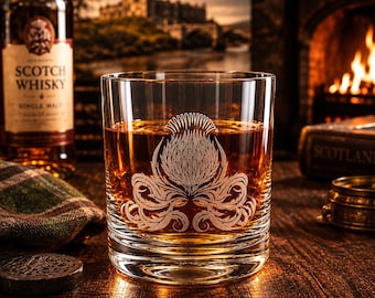 May include: A clear glass tumbler filled with amber liquid, featuring an etched thistle design on the front. The glass has a thick base and horizontal ridges near the bottom, holding a golden-brown spirit.