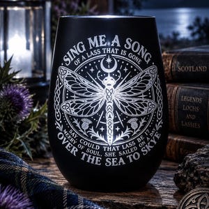 Könnte beinhalten: Ein schwarzes, stieloses Weinglas mit einer weißen Gravur, die eine Libelle, eine Mondsichel und Distelblüten zeigt. Der Text lautet: SING ME A SONG OF A LASS THAT IS GONE, SAY, COULD THAT LASS BE I? MERRY OF SOUL, SHE SAILED ON A DAY OVER THE SEA TO SKYE.