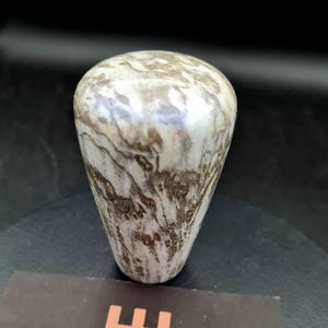 Könnte beinhalten: Ein Schaltknauf mit einer glatten, polierten Oberfläche, die ein wirbelndes, marmoriertes Muster in Weiß- und Erdbrauntönen aufweist. Das Objekt hat eine konische, zylindrische Form mit abgerundeter Oberseite und einer steinartigen Textur.