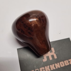 Puede incluir: Un pomo de palanca de cambios de madera brillante, en forma de lágrima, de color marrón oscuro. La superficie es lisa y pulida, con vetas de madera visibles. Una tarjeta de visita con el texto "JACKKNOBZ" está en el fondo.