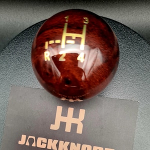 Puede incluir: Un pomo de palanca de cambios esférico con un acabado de grano de madera marrón oscuro pulido. La superficie superior presenta un patrón de cambio manual dorado que muestra las marchas 1 a 4 y R. Debajo del pomo hay una tarjeta negra con el logo JACKKNOB.