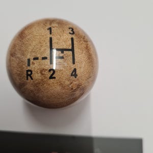 May include: A wooden gear shift knob with a light brown finish. The shift pattern is printed in black, with numbers and an "R" for reverse. The knob is spherical and sits on a white surface.