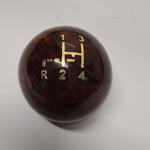 Può includere: Un pomello del cambio sferico in legno marrone scuro con finitura lucida. La superficie superiore presenta un inserto color oro che mostra lo schema di una trasmissione manuale a quattro velocità con i numeri 1, 2, 3, 4 e la lettera R.