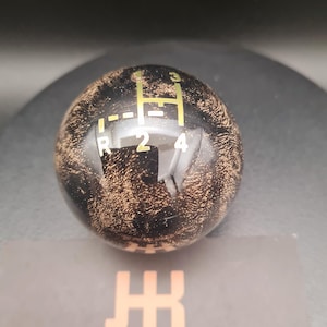 Op de afbeelding: Een bolvormige versnellingspookknop met een zwart en koperkleurige marmeren afwerking. Het bovenoppervlak is voorzien van een goudkleurig versnellingspatroon met de cijfers 1, 2, 3, 4 en R. Onder de knop ligt een zwarte kaart met het logo JK en JACKKNOBZ.